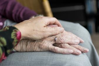 Two hands of one elderly person in their lap with another elderly persons hand on top.